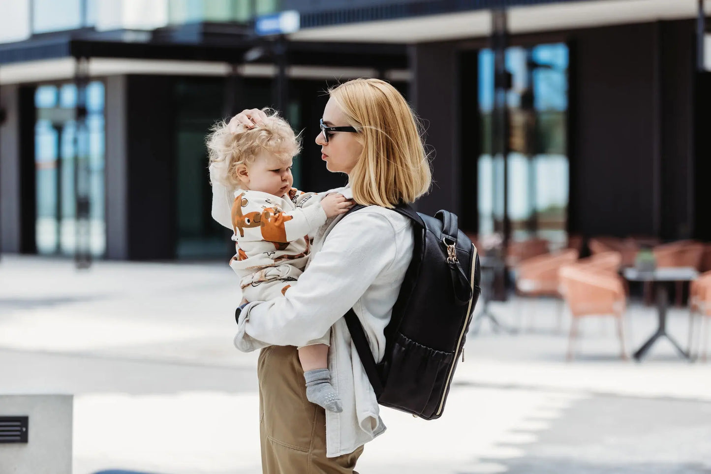 A woman holding a baby in a stylish Espresso Diaper Bag, showcasing its spacious design with 16 pockets, laptop and baby food compartments, stroller straps, and waterproof changing mat.