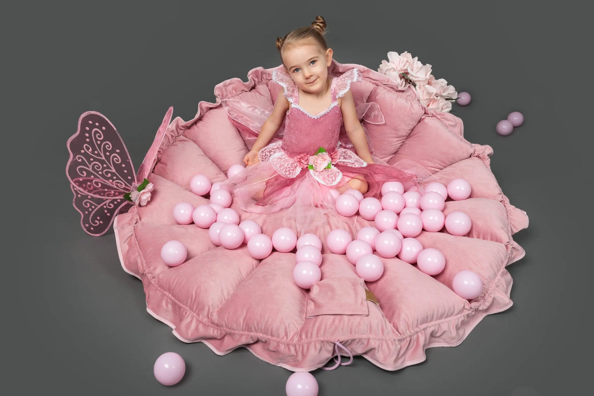 A girl in a pink dress plays with a flower-shaped playmat transforming into a ball pit, surrounded by pink balls. Includes 100 balls for motor and sensory development.