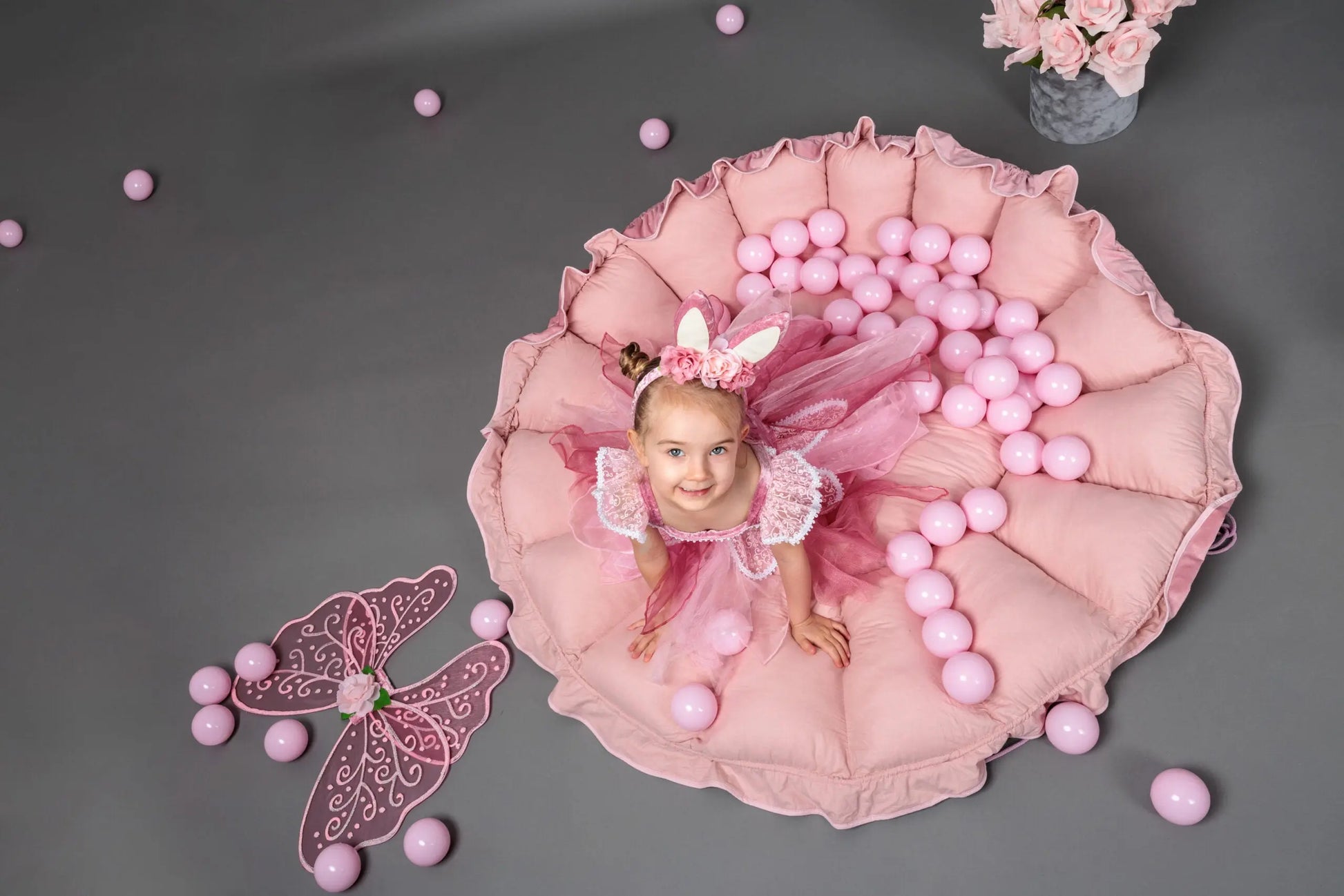 A girl in a pink dress playing in a Flower Play Mat with Ball Pit, surrounded by pink butterfly wings and balls, enhancing sensory development and imaginative play.