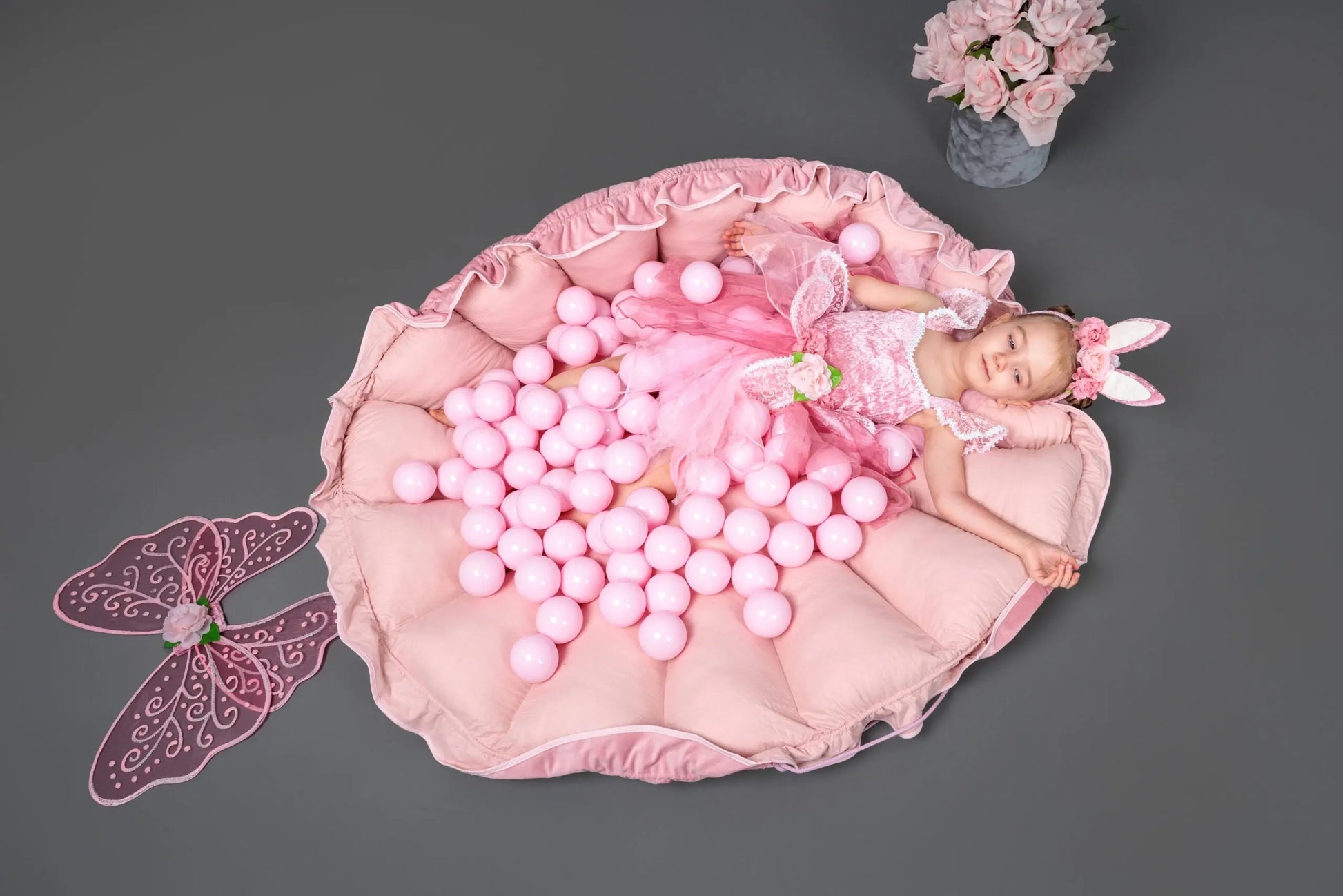 A girl in a pink dress lies on a pink pillow with pink balls, showcasing a Flower Play Mat with Ball Pit - Grey and White, a versatile 2-in-1 set for children's play and development.