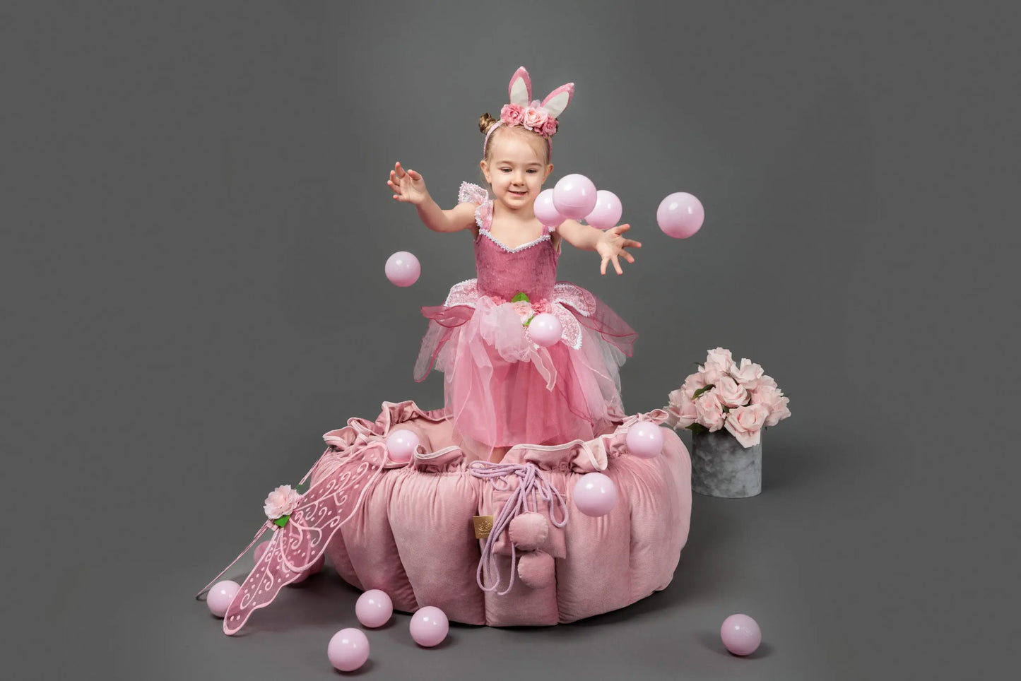 A girl in a pink dress juggling balls on a flower playmat transforming into a ball pit, surrounded by a delicate ruffle. 100 balls included for motor and sensory development.