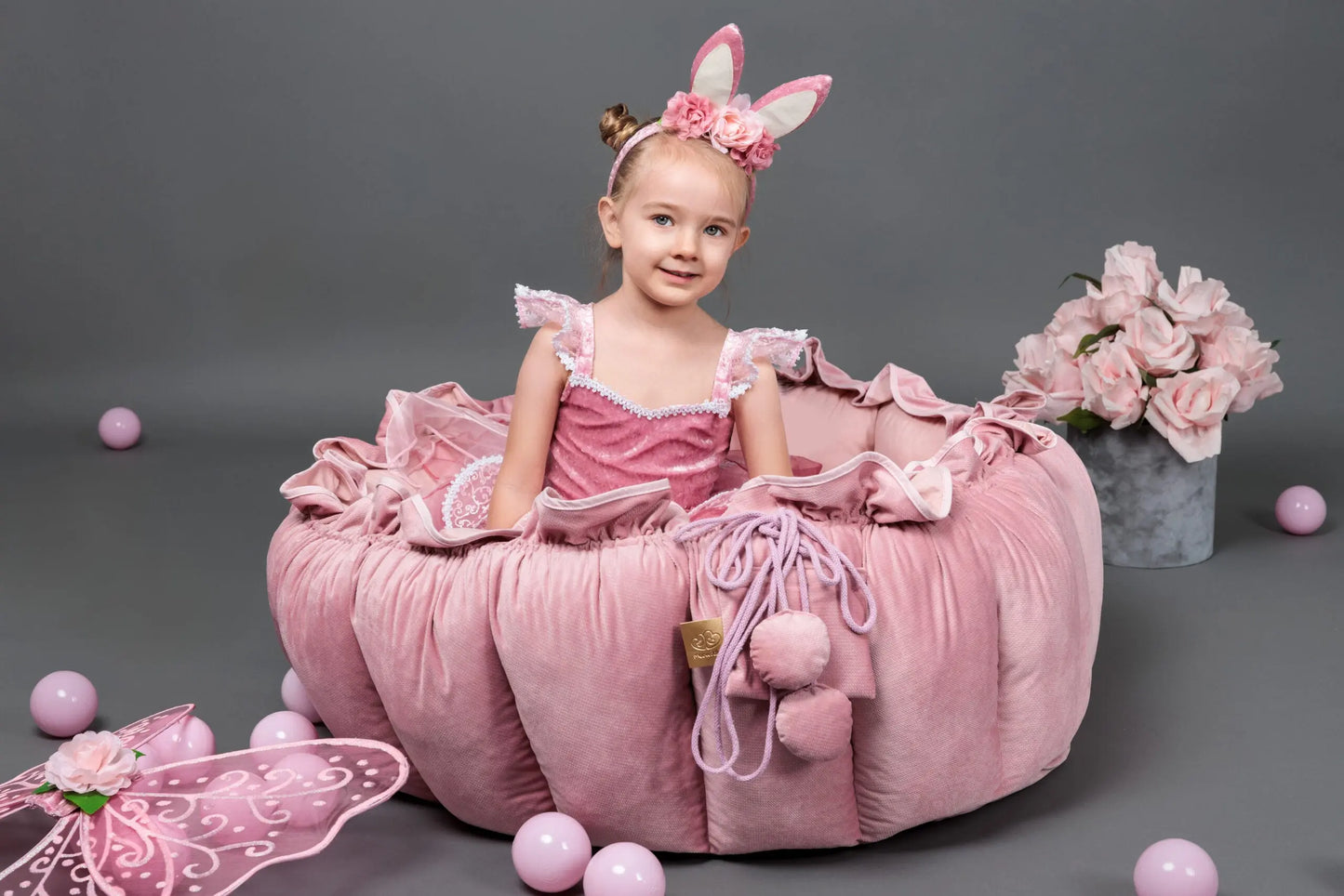 A girl in a pink dress plays with a flower-inspired playmat, transforming into a ball pit. Set includes 100 balls for sensory and motor development. Made of soft cotton and velvet.