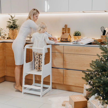 A child stands on a Montessori Learning Tower Step Stool in a kitchen, promoting independence and safe exploration. Handmade from Baltic birch plywood, height-adjustable for young learners.