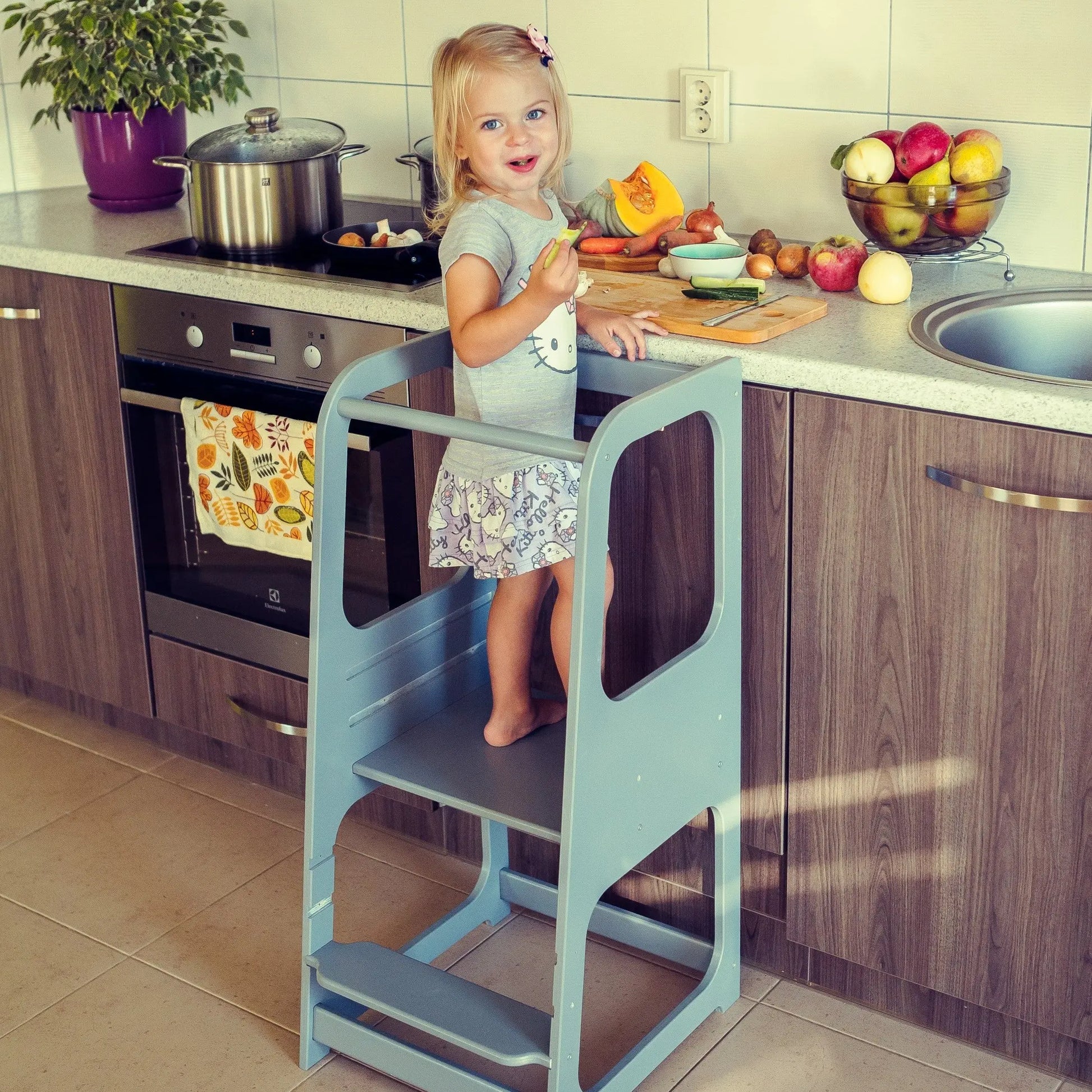 A child stands on a Montessori Helper Tower Step Stool in a kitchen, promoting independence and safe interaction. Handmade from Baltic birch plywood, height-adjustable for young learners.