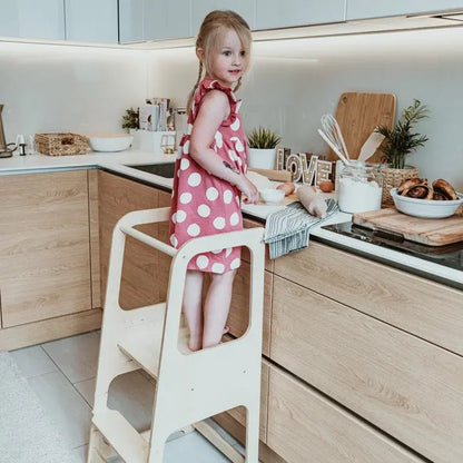 A child stands on a Montessori Learning Tower Step Stool in a kitchen, promoting independence and safe interaction. Adjustable height, sturdy Baltic birch plywood construction, and handmade quality from Latvia.