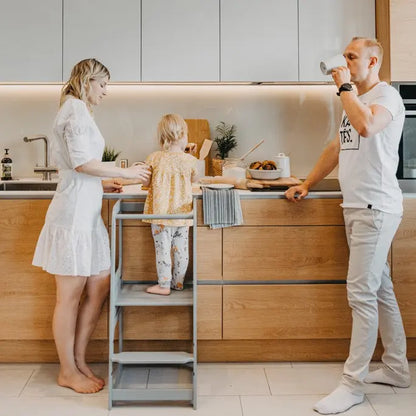 A man and woman with a child in a kitchen, showcasing the Montessori Learning Tower Step Stool for Kids. Height-adjustable design, sturdy Baltic birch plywood construction. Encourages independence and safe exploration in the kitchen.