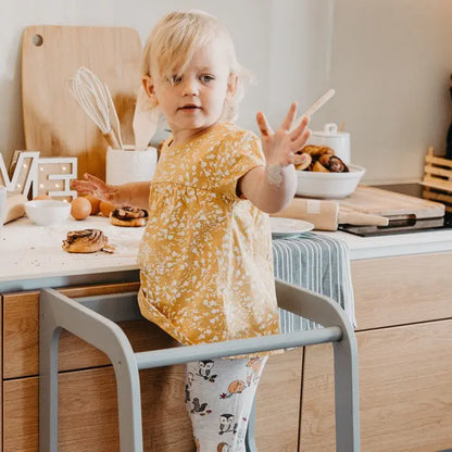 A child stands in a Montessori Learning Tower Step Stool, hands raised. Height-adjustable design for kitchen or bathroom use, encouraging independence and safe exploration. Made from sturdy Baltic birch plywood.