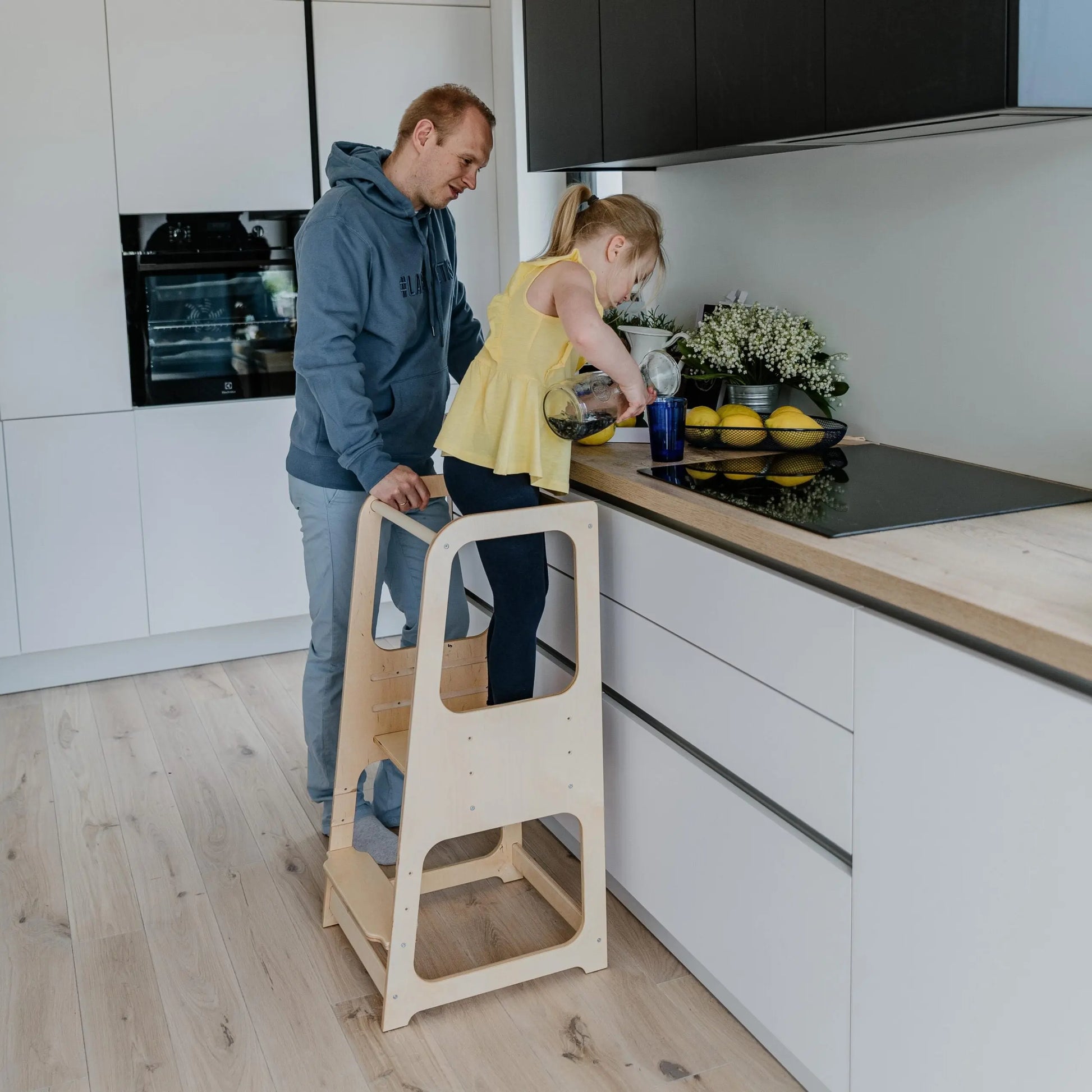 A man and child stand on a Montessori Learning Tower Step Stool in a kitchen. Height-adjustable design for kids, made from sturdy wood, encouraging independence and safe kitchen exploration.