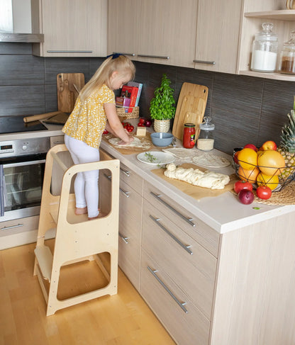A child stands on a Montessori Learning Tower Step Stool in a kitchen, reaching for items. Adjustable height, sturdy Baltic birch plywood construction. Encourages independence and safe exploration.