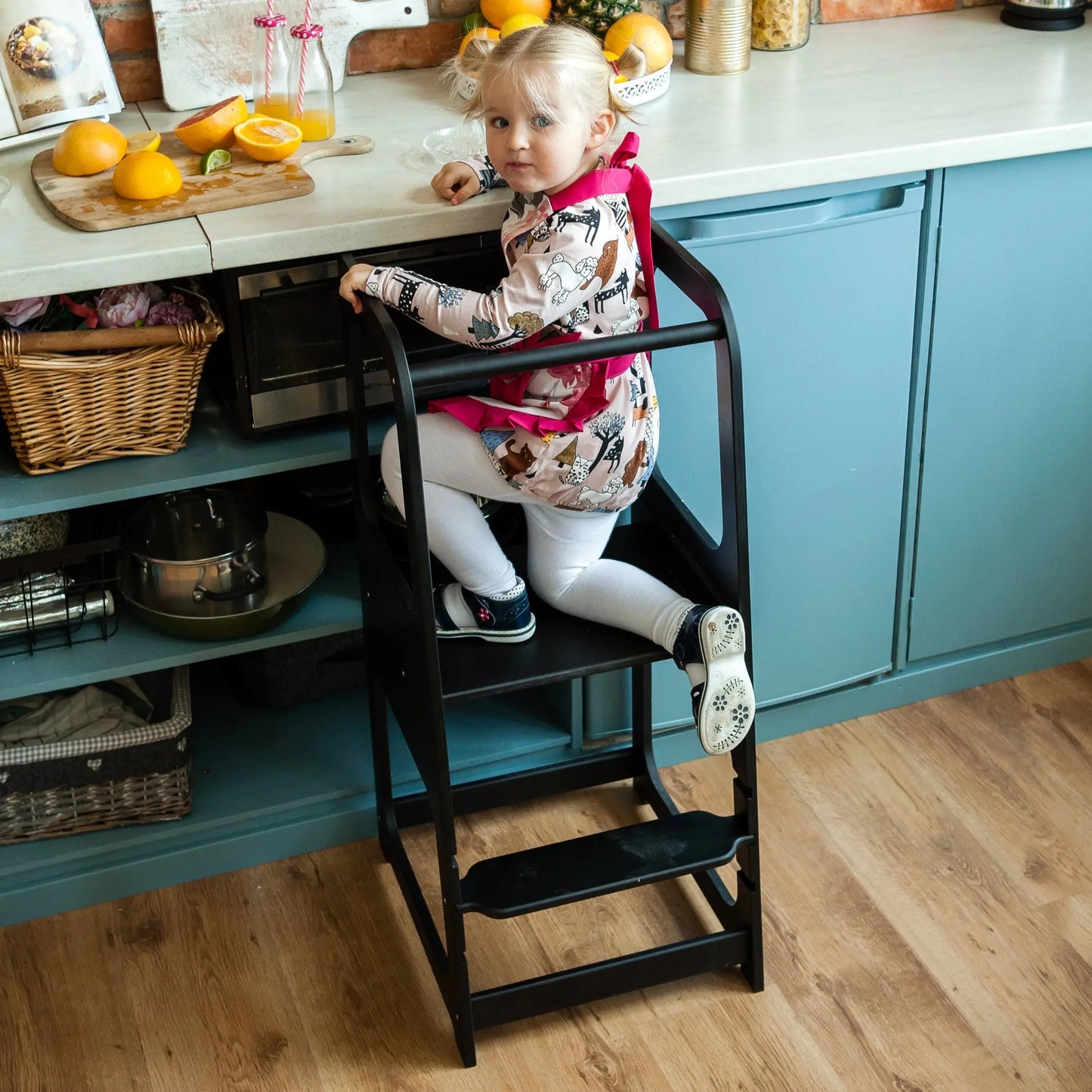 A child sits on a Montessori Learning Tower Step Stool in a kitchen, reaching for items. Adjustable height, sturdy Baltic birch plywood, handmade in Latvia. Encourages independence and safe kitchen interaction.