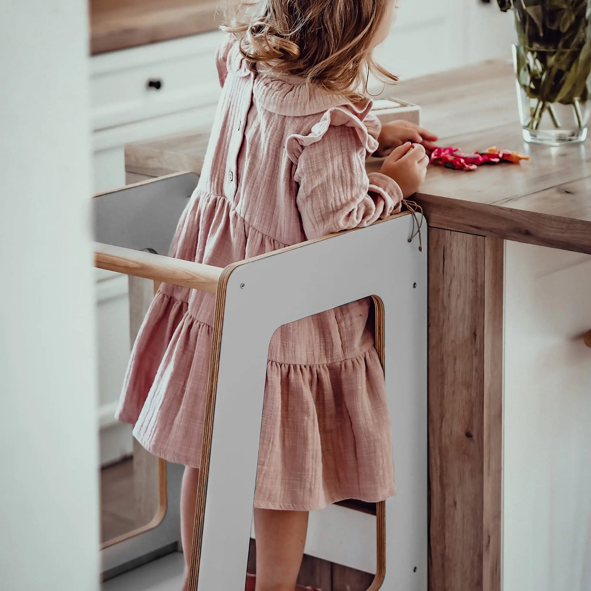 A girl in a pink dress stands at a table, using the Plywood Kitchen Helper Scandi. Made of birch plywood with adjustable platform heights, it promotes kitchen safety and independence for children.