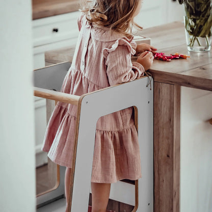 A girl in a pink dress stands at a table, using the Plywood Kitchen Helper Scandi. Made of birch plywood with adjustable platform heights, it promotes kitchen safety and independence for children.