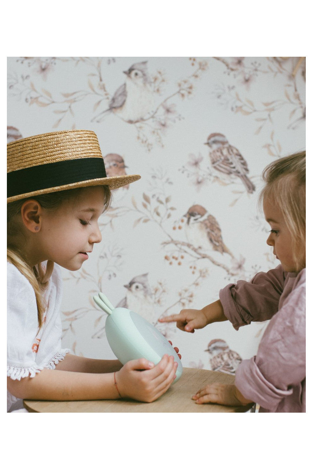 A girl in a hat points at a small round object, showcasing the Rabbit Lamp with Alarm Clock - Green. Features sunrise and sunset functions, nature sounds, touch control, and more.