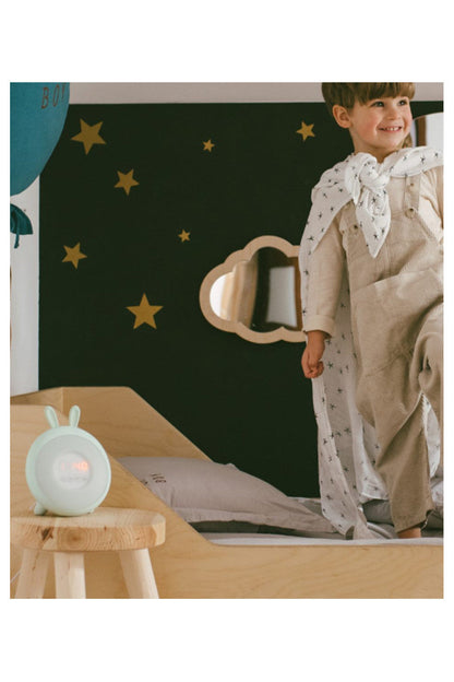 A boy standing on a bed, a white alarm clock with ears, a clock on a stool, and a person holding a pillow. Product: Rabbit Lamp with Alarm Clock - Green. Mimics sunrise and sunset light intensities, aiding in healthy sleep routines for all ages.