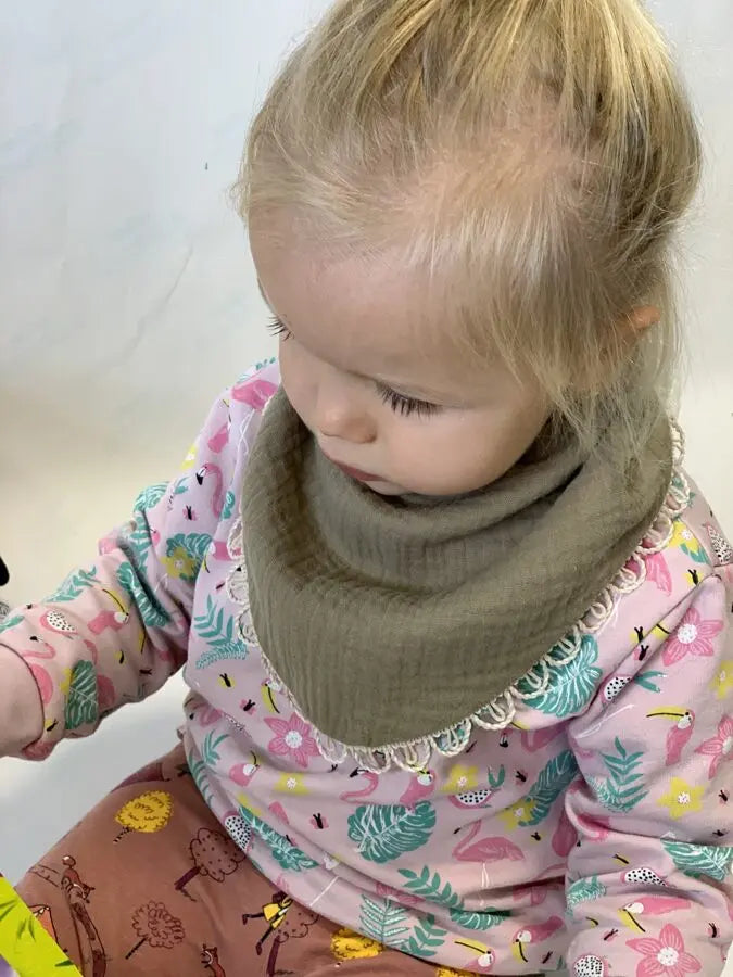 A child in a khaki Scarf-Bib, holding a pen, with a close-up of their face. Handmade muslin fabric, soft and protective for ages 6 months to 3 years.