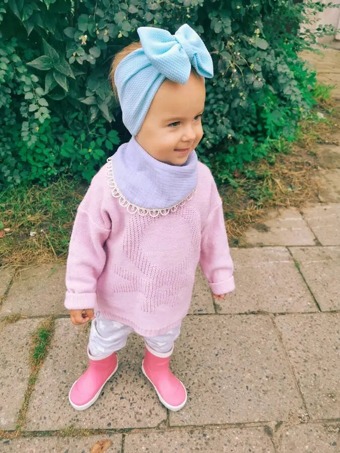 A child in a pink sweater and blue hat wears a Scarf-Bib - Light Blue, made of 100% muslin fabric for softness and style. Suitable for ages 6 months to 3 years.