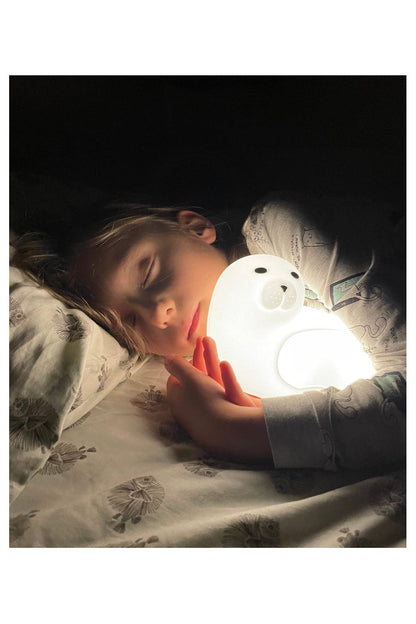 A girl sleeping with a seal-shaped lamp emitting warm light, featuring a playful seal design with a remote control. Soft, safe silicone material, touch-responsive, 7-color lighting, USB-C charging, and 2-hour auto-off.