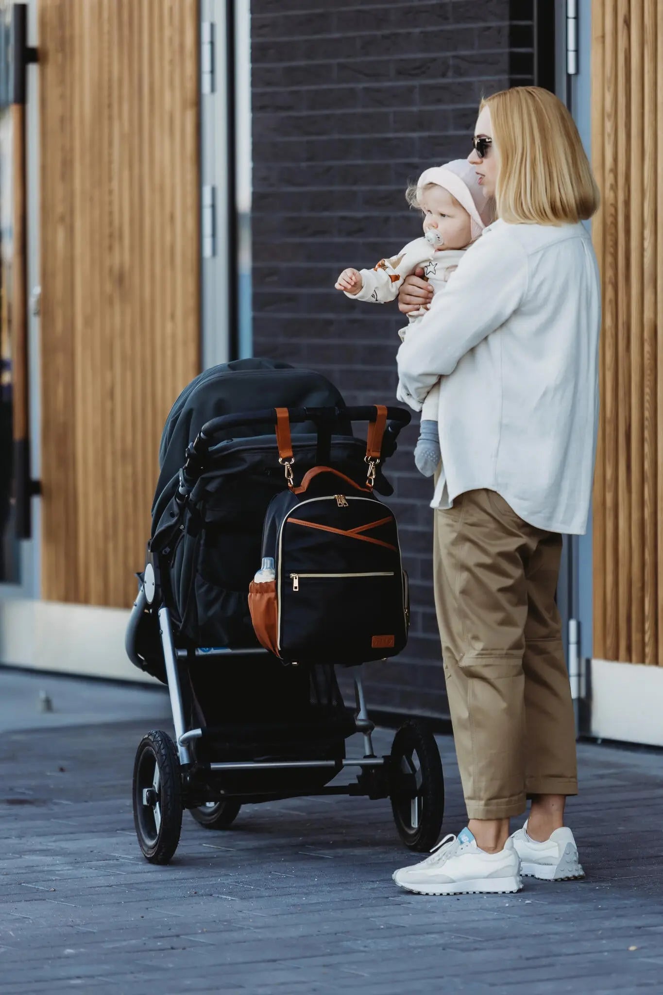 A woman holds a baby near a black and brown backpack on a stroller. Small Diaper Backpack – Black Coffee by Ally Scandic, designed for style, organization, and convenience on the go.