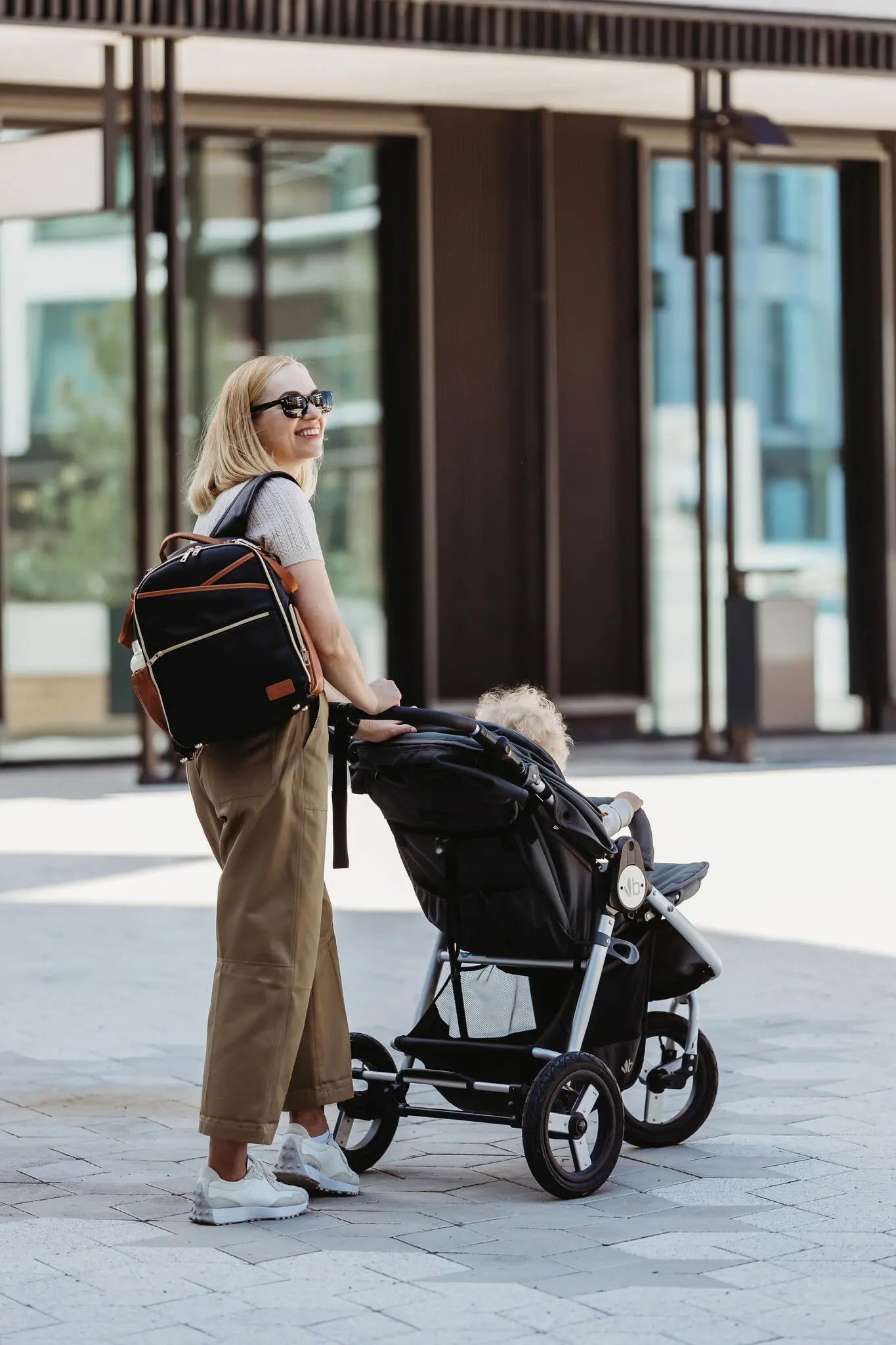 A woman pushing a stroller with a baby, carrying a backpack and bag. Small Diaper Backpack – Black Coffee by Ally Scandic, designed for style, organization, and convenience on the go.