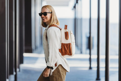 A woman wearing sunglasses and a backpack, showcasing the Small Diaper Backpack – Cappuccino by Ally Scandic. Stylish, lightweight design with multiple pockets and stroller straps for on-the-go parents.