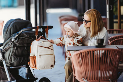 A woman and baby admire the Small Diaper Backpack in Cappuccino. Stylish, lightweight PU vegan leather with multiple pockets and stroller straps for hands-free convenience. Dimensions 35 × 26 × 15 cm.