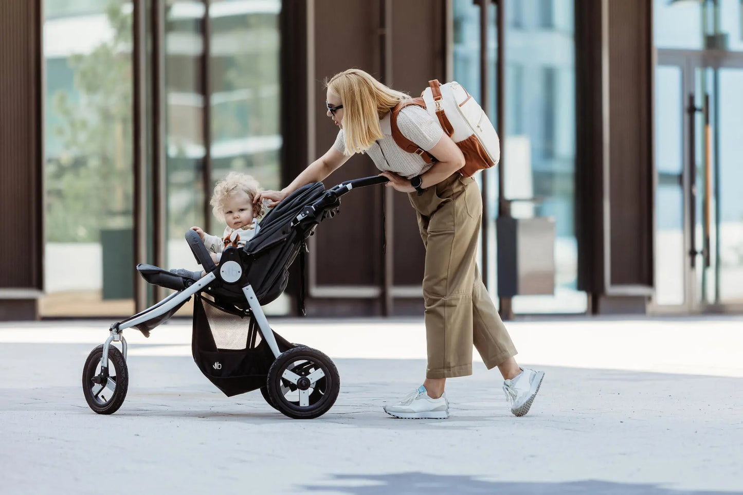 A woman with blonde hair pushing a Cappuccino Diaper Bag stroller. Lightweight, versatile design with multiple pockets, laptop compartment, and stroller straps for hands-free convenience.