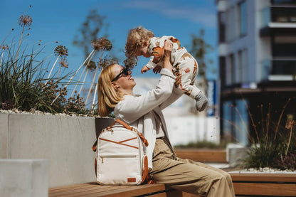 A woman holds a baby while wearing a white and brown Small Diaper Backpack – Cappuccino outdoors. The backpack features multiple pockets, thermal linings, and stroller straps for convenient parenting on the go.