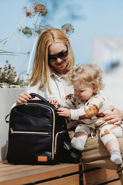 A woman holds a baby, examining a Small Diaper Backpack – Espresso. Lightweight, vegan leather, multiple pockets, and stroller straps for hands-free convenience. Functional and stylish for on-the-go parents.