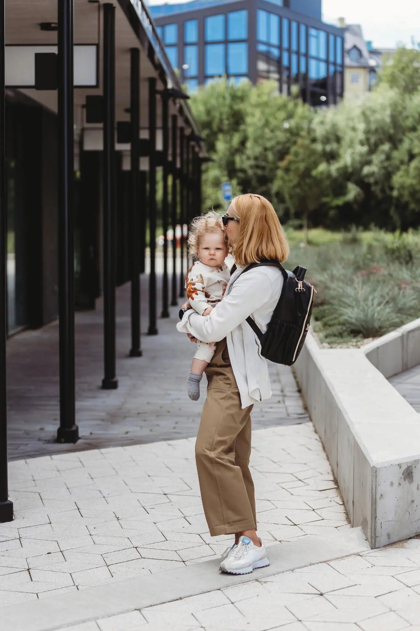 A woman carrying a Small Diaper Backpack in Espresso, featuring multiple pockets, stroller straps, and durable vegan leather, designed for style and convenience by Ally Scandic.