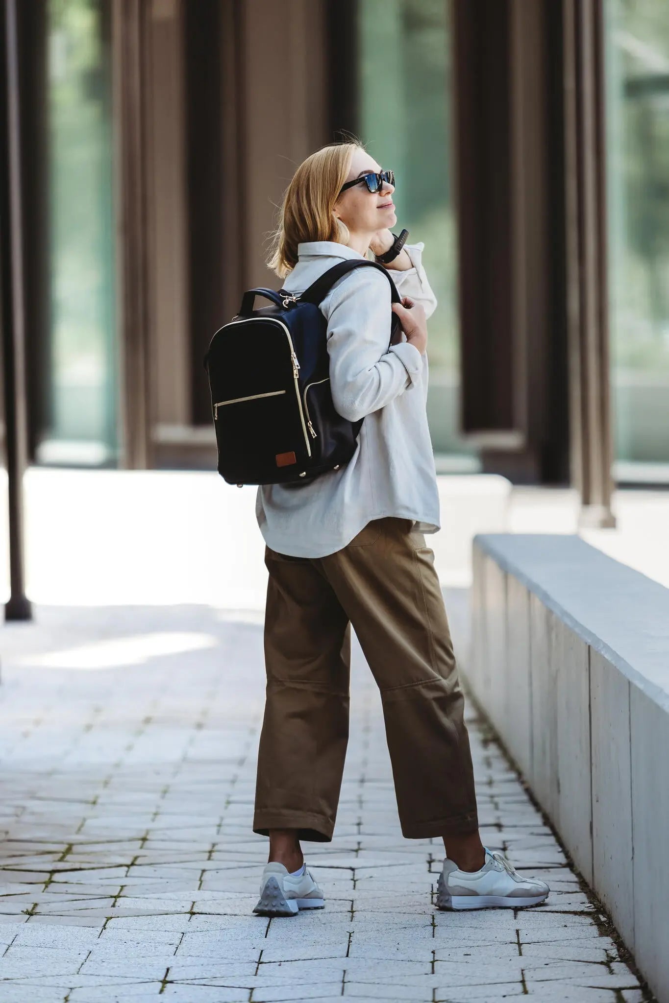 A woman in street fashion wears a Small Diaper Backpack – Espresso. Lightweight, vegan leather with multiple pockets for organized essentials, perfect for stylish, on-the-go parents by Ally Scandic.