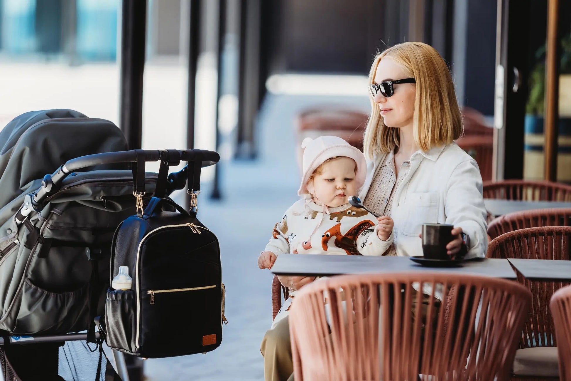 A woman holding a baby with a black backpack on a stroller, showcasing the Small Diaper Backpack in Espresso. Designed for style and convenience, featuring multiple pockets and stroller straps for hands-free use.