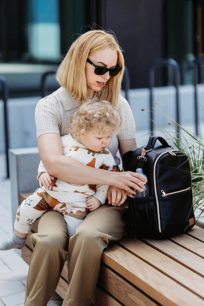 A woman with blonde hair holds a baby while wearing sunglasses, showcasing the Small Diaper Backpack in Espresso by Ally Scandic for stylish and organized parenting on the go.