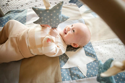 A baby lies on a starry blanket with interactive arches and toys - rattle star, rustling star, and rattle star. Handmade 80x80 cm activity mat in calming blue for sensory exploration.