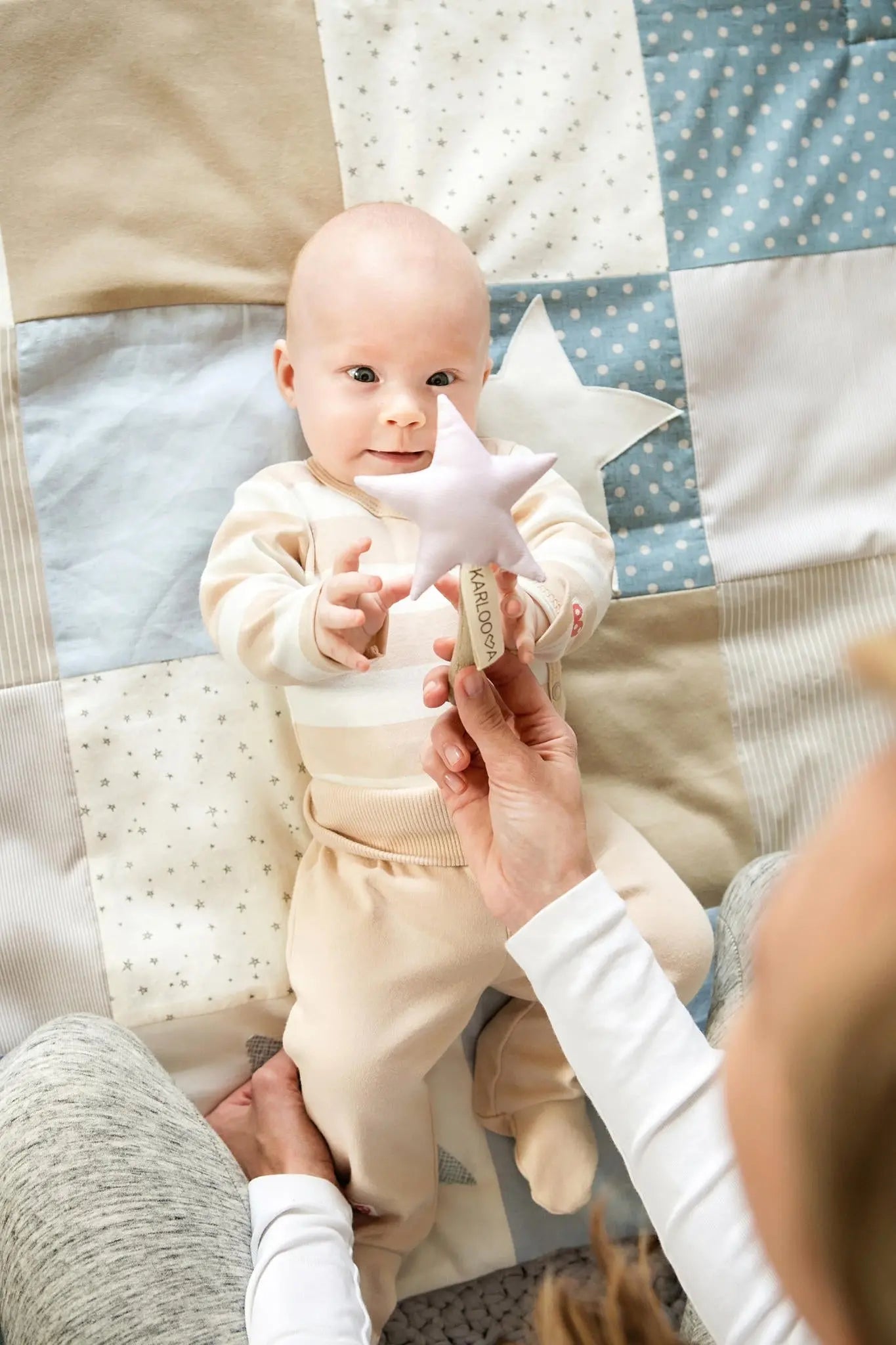 A baby lies on a blue activity mat with interactive arches, holding a star-shaped toy. Mat features detachable arches, rattle stars, and sensory stimulation for newborns.