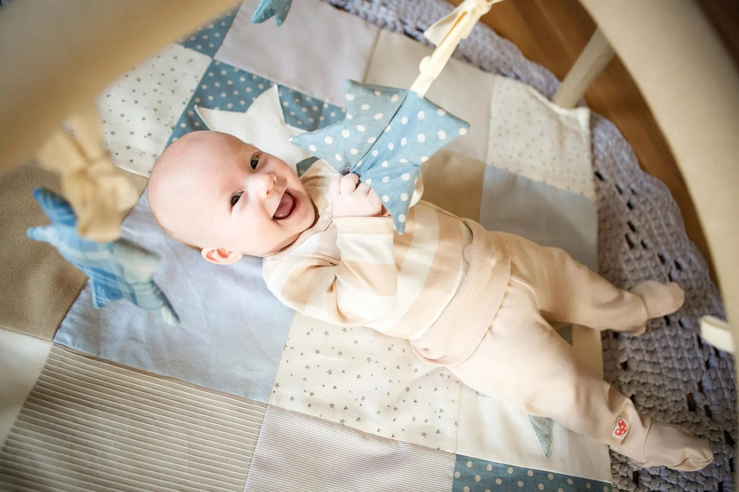 A baby lies on an activity mat holding star-shaped toys under interactive arches. Mat stimulates visual and motor skills with contrasting colors and detachable arches. Crafted from ecologic cotton.