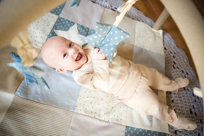 A baby lies on an activity mat holding star-shaped toys under interactive arches. Mat stimulates visual and motor skills with contrasting colors and detachable arches. Crafted from ecologic cotton.