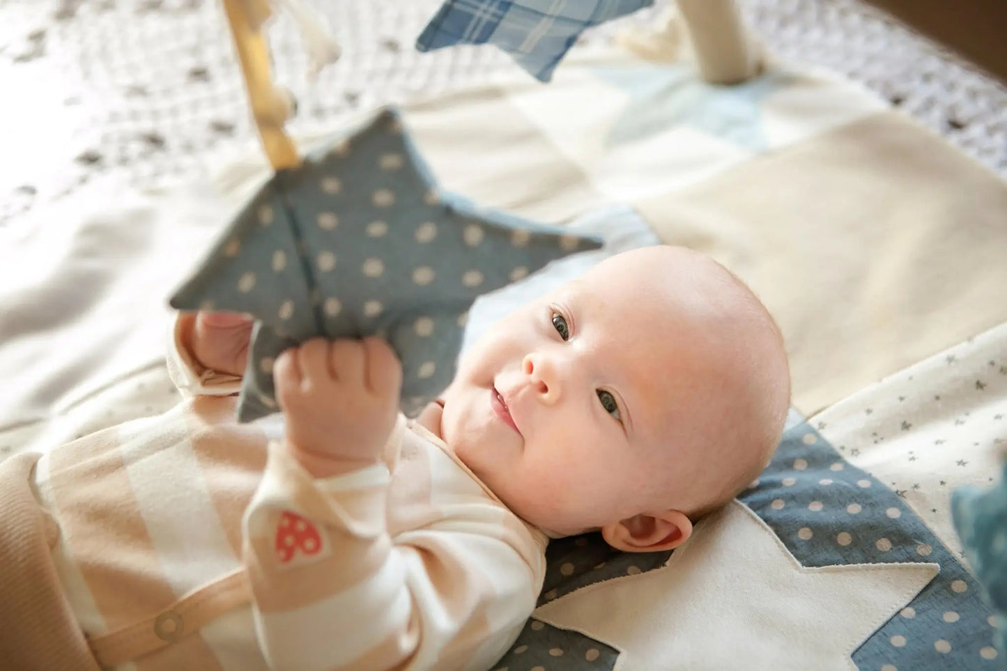 A baby lies on an activity mat, holding a star toy, surrounded by sensory arches. Mat features contrasting colors, detachable arches with sounds, and eco-friendly cotton material.