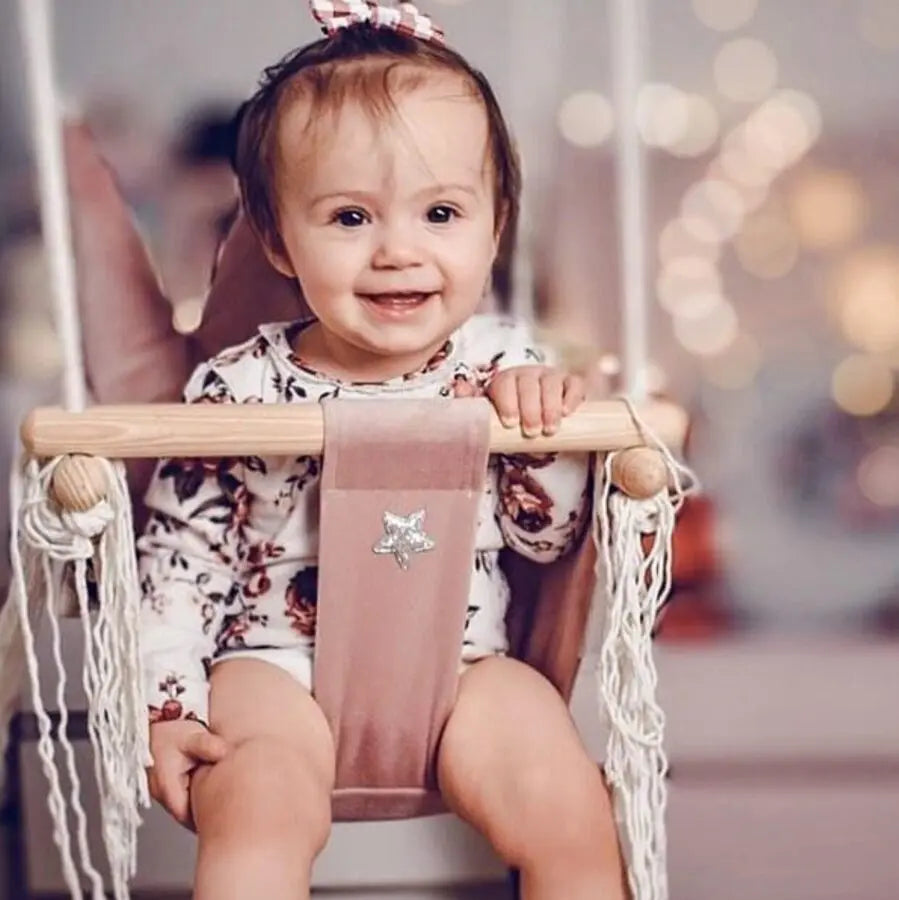 A toddler happily swings on the Crown Shaped Cushion Swing in Dusty Pink, featuring soft velvet plush, durable pinewood, and a podium crown for comfort.