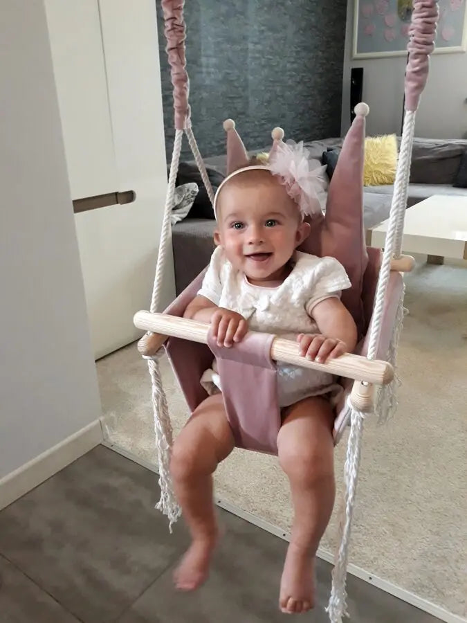 A toddler joyfully swings in a Crown Shaped Cushion Swing. Soft velvet plush material, sturdy pinewood construction, and a podium crown for comfort. Dusty Pink.