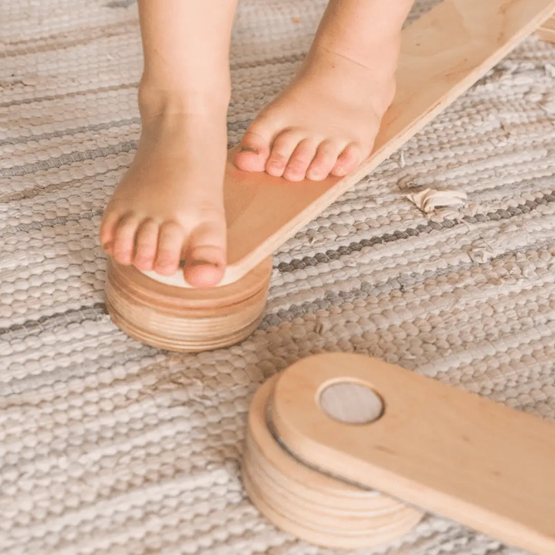 A child's bare feet on a wooden balance beam, promoting balance and coordination. Crafted from safe plywood, includes connectors for assembly. Enhance motor skills with our Wooden Balance Beams.