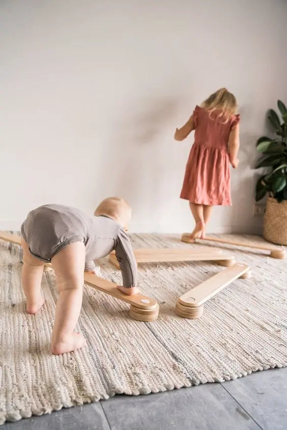 A baby playing on Wooden Balance Beams, aiding in gross motor skills and balance control. Set includes three beams and connectors, promoting creativity and safe play. Made in Europe for quality assurance.