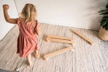 A girl in a pink dress plays on Wooden Balance Beams, aiding in self-confidence and motor skills development. Set includes beams and connectors for up to 50 kg load capacity.