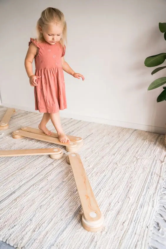 A toddler in a pink dress stands on a Wooden Balance Beam, pointing at her dress. Wooden planks and a leaf close-up are visible. Product promotes balance, motor skills, and creativity.