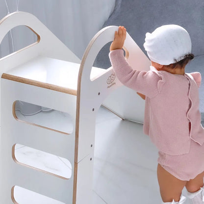 Toddler in pink outfit standing on Wooden Indoor Scandi Slide, 87x46cm, with white edges. Slide features stable structure, 3 steps, and handles for safe climbing. Ideal for kids over 12 months.