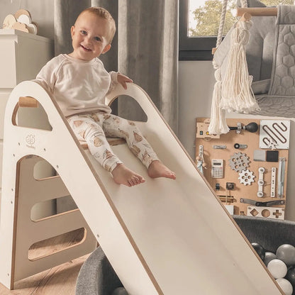 Child on a wooden indoor Scandi slide, 87x46cm, with three steps, smooth edges, and comfortable handles for safe play. Made of birch plywood, supporting motor skills and coordination.