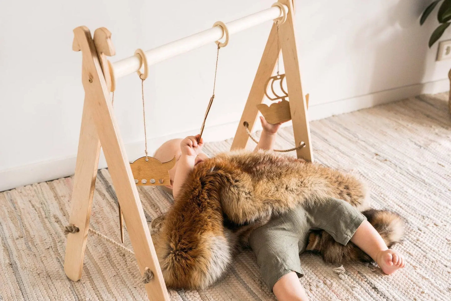 A baby lies on a rug under a wooden play gym with Viking-themed hanging toys. Gym promotes muscle development and coordination for ages 0-12 months.