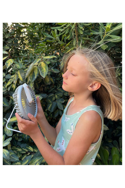 A girl holding a small fan with adjustable air flow and built-in lamp. Portable and rechargeable, perfect for hot days and nights. Dimensions: 15.4 x 14.5 x 7.6 cm.