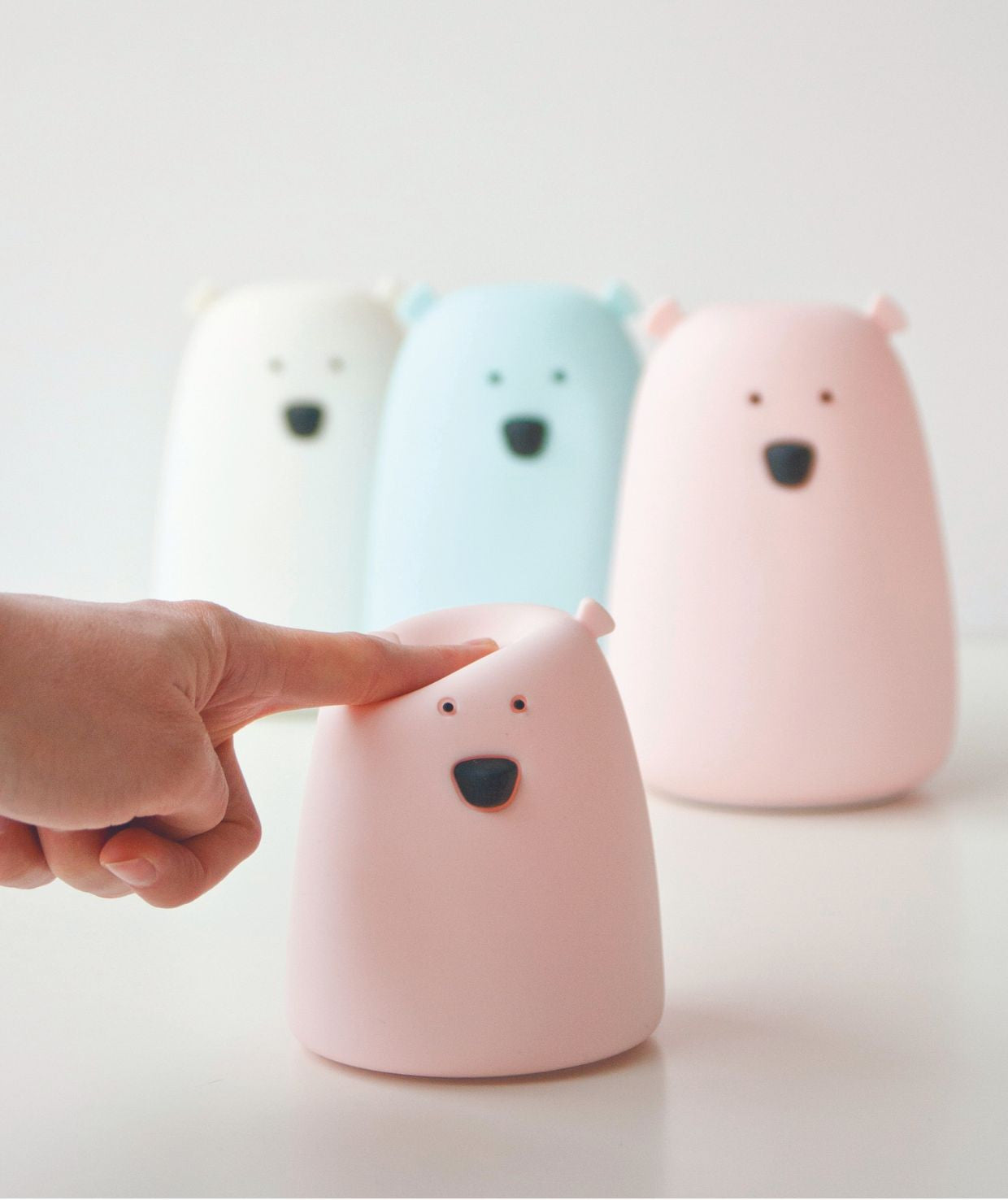 A finger points at a pink silicone lamp shaped like a friendly bear face. Soft, touch-operated night light in 7 colors, safe for kids, with 12-hour battery life.