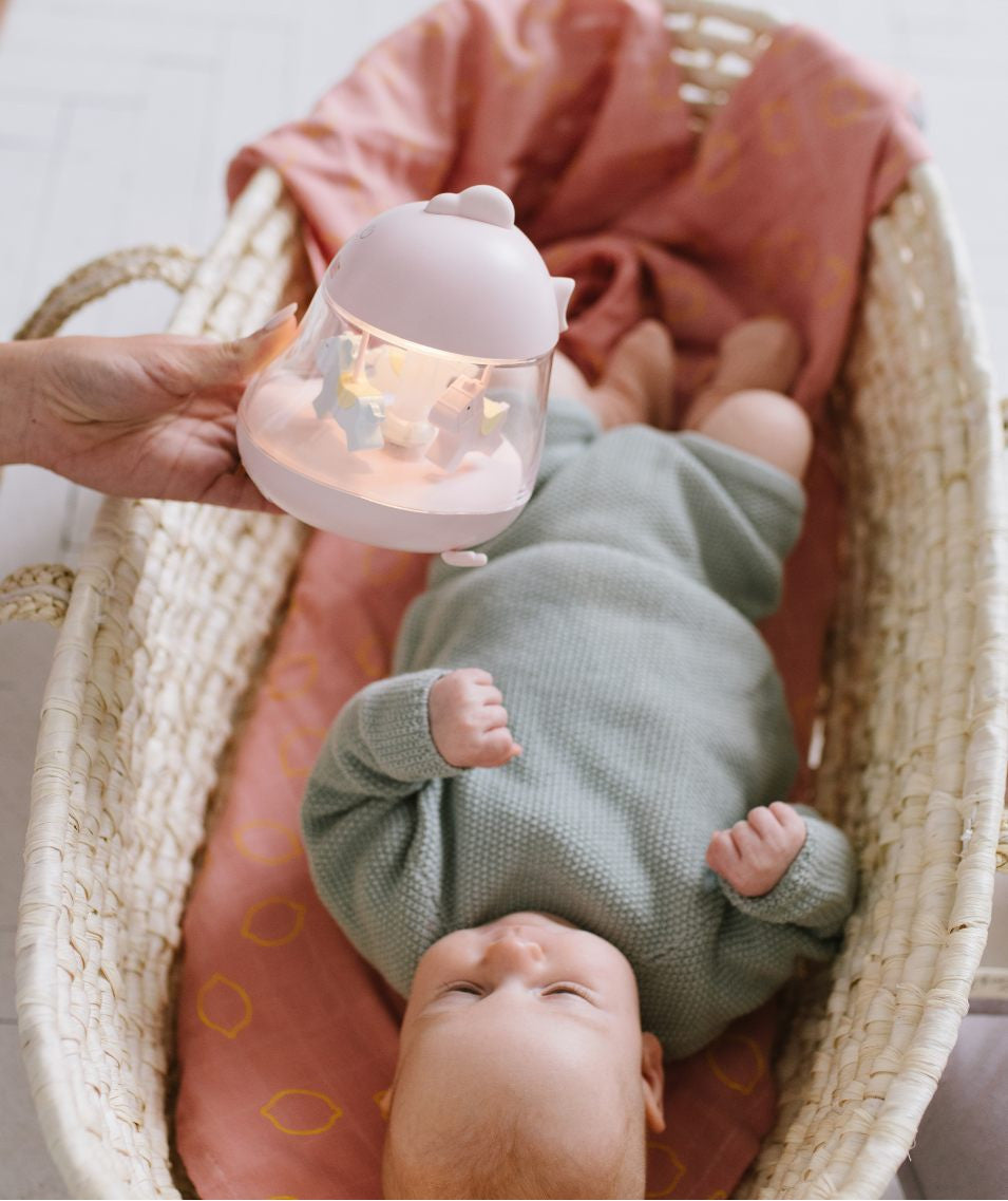 A baby lying in a crib with a lamp featuring a carousel and music box. Carousel horses spin, soothing melody plays, and the lamp illuminates in 7 colors. Dimensions: 15.4 x 12.6 x 11.4 cm.