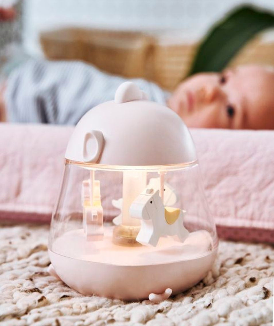 A baby lies on a blanket near a white carousel with horses. Close-ups show the baby's head and face, and a white object. The product is a Lamp with Carousel and Music - Pink, featuring a merry-go-round design, music box melody, color-changing light, and USB charging.